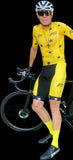 Person in yellow cycling outfit with helmet and sunglasses standing next to a bicycle on a black background